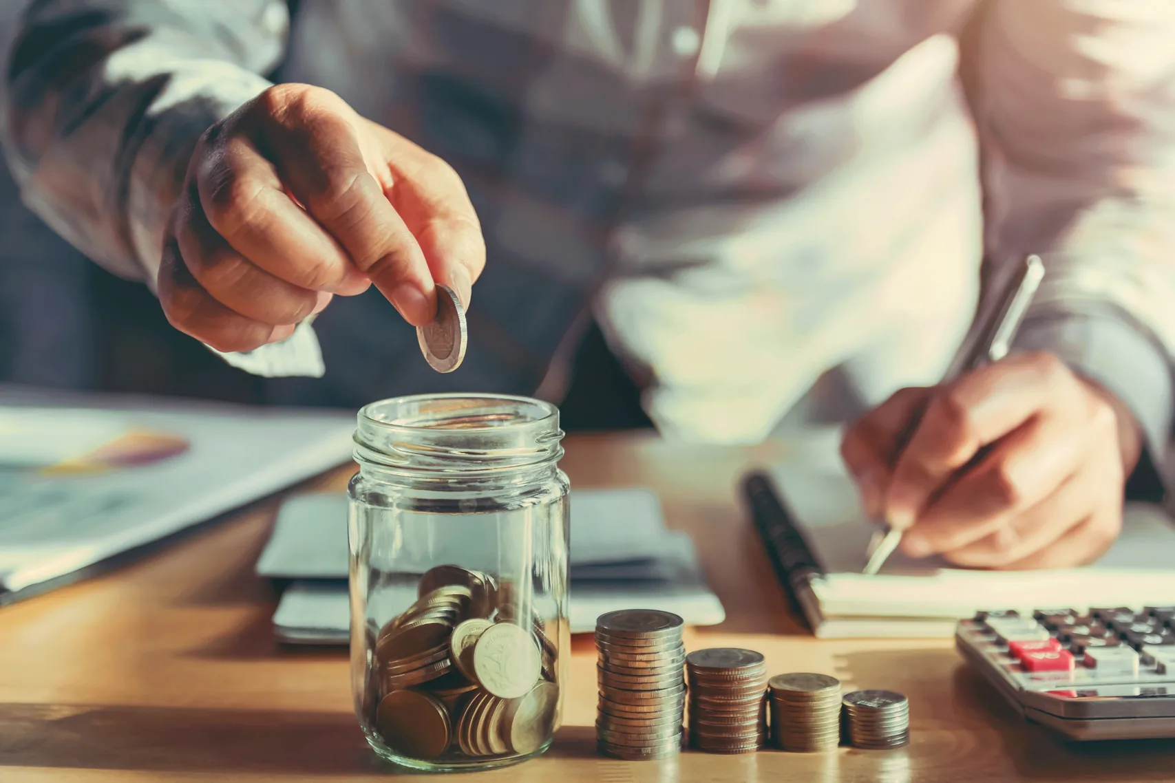 A hand drops a coin into a glass jar beside stacks of coins and a calculator