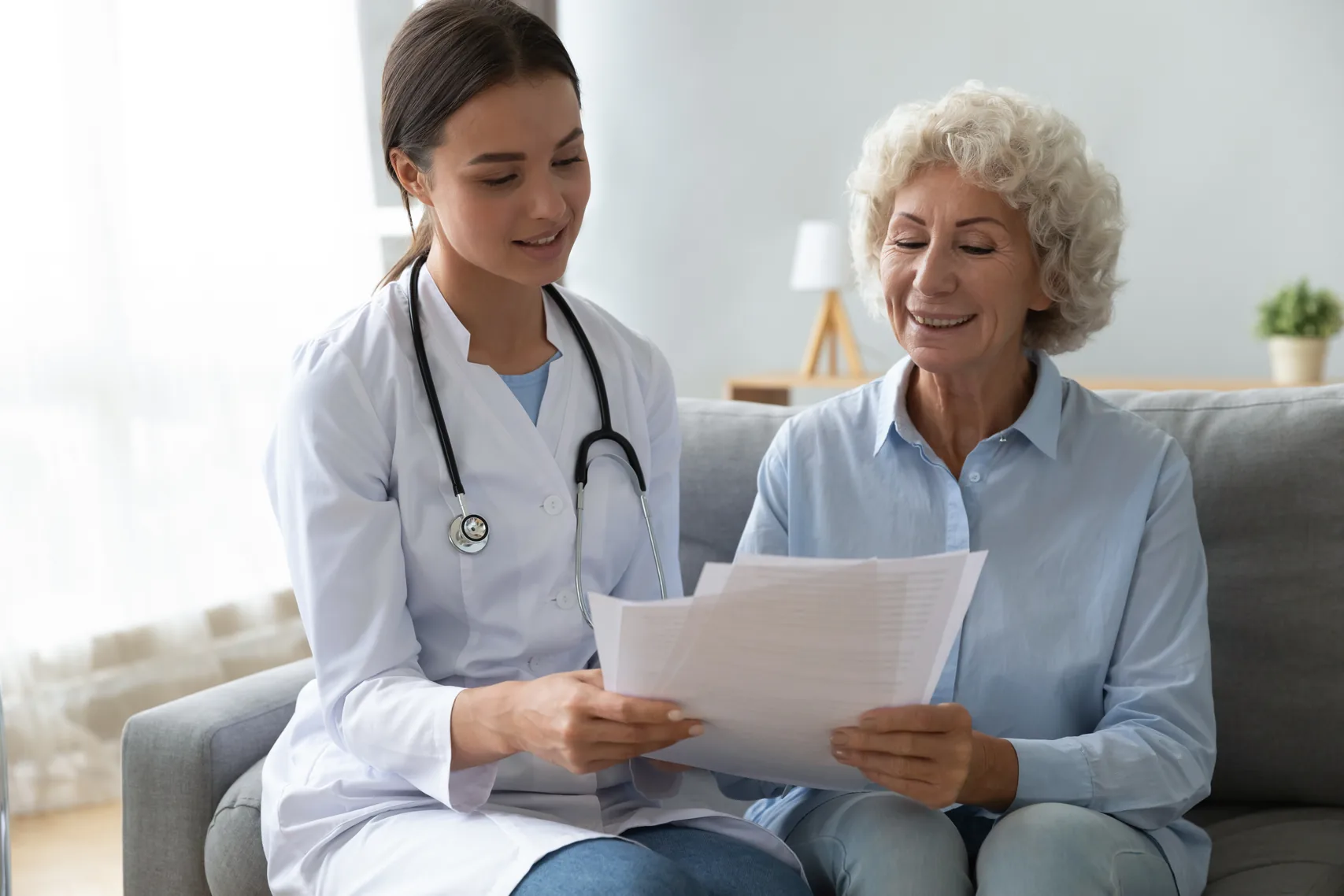 A young doctor goes over paperwork with an elderly patient on a sofa