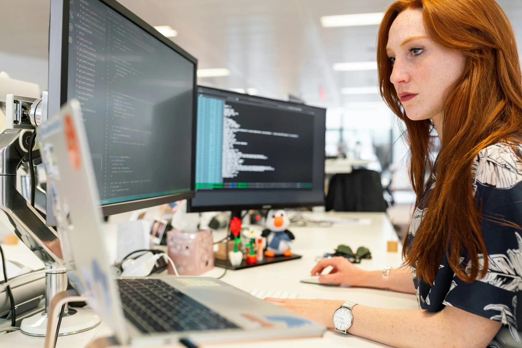 A red-haired woman works at a desk with two monitors showing code
