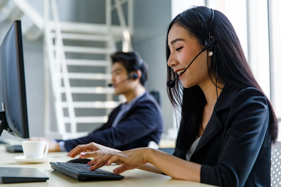 A smiling call center agent in a headset types at her desk, with a colleague in a headset behind her