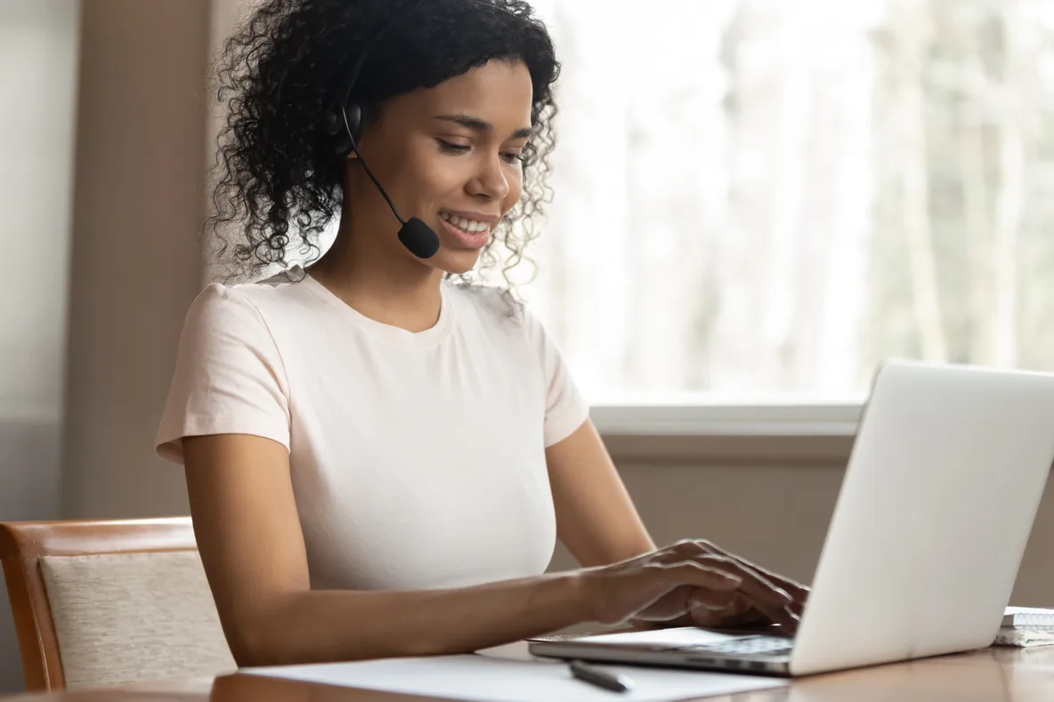 A smiling transcriptionist wearing a headset types on a laptop